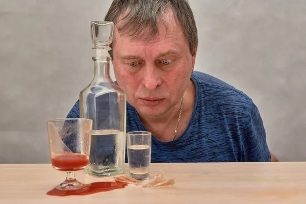 Een trieste man zit aan een tafel en drinkt alleen wodka met alcoholverslaving.. — Stockfoto