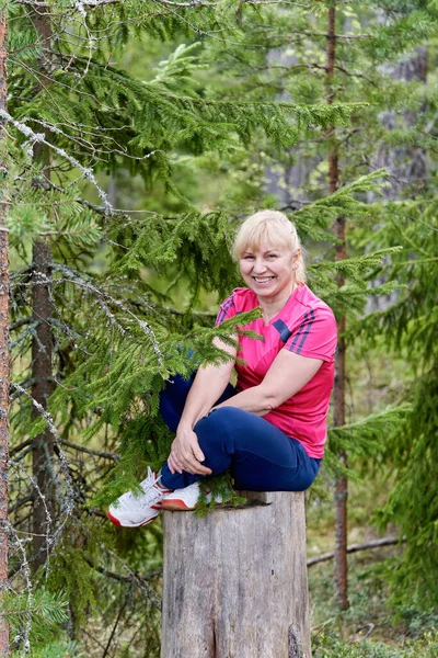 Женщина средних лет со светлыми волосами сидит на высокой пне в лесу — стоковое фото
