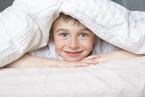 Χαμογελαστό αγόρι που κρύβεται στο κρεβάτι κάτω από μια κουβέρτα λευκό ή ημίδιπλο βαμβακερό. — Φωτογραφία Αρχείου