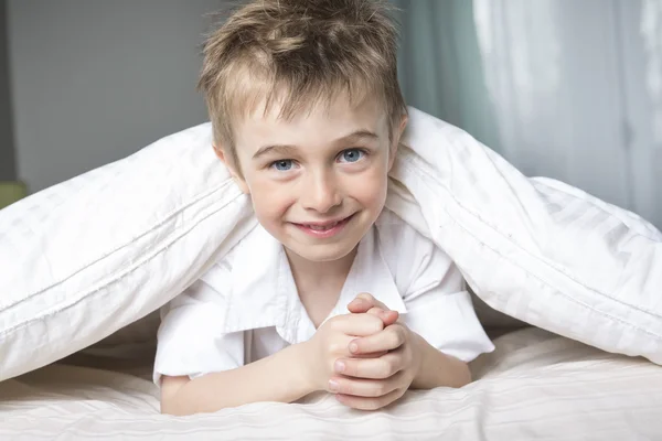 Garoto sorridente escondido na cama sob um cobertor branco ou um cobertor . — Fotografia de Stock