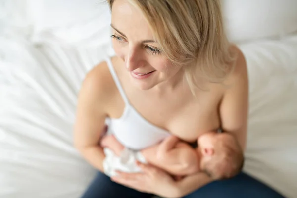 Mujer en su dormitorio en los cojines blancos, sonriendo y amamantando a su bebé. — Foto de Stock