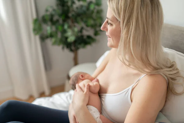 Mulher em seu quarto nas almofadas brancas, sorrindo e amamentando seu bebê. — Fotografia de Stock