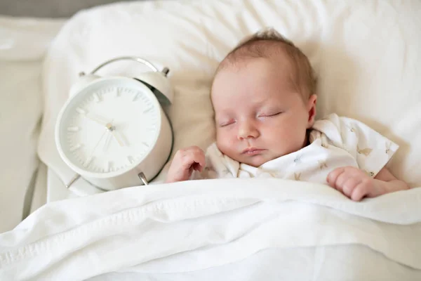 Ένα γλυκό νεογέννητο κοριτσάκι κοιμάται στο λευκό κρεβάτι με το ρολόι στο πλάι — Φωτογραφία Αρχείου