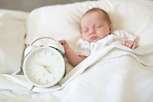Ένα γλυκό νεογέννητο κοριτσάκι κοιμάται στο λευκό κρεβάτι με το ρολόι στο πλάι — Φωτογραφία Αρχείου