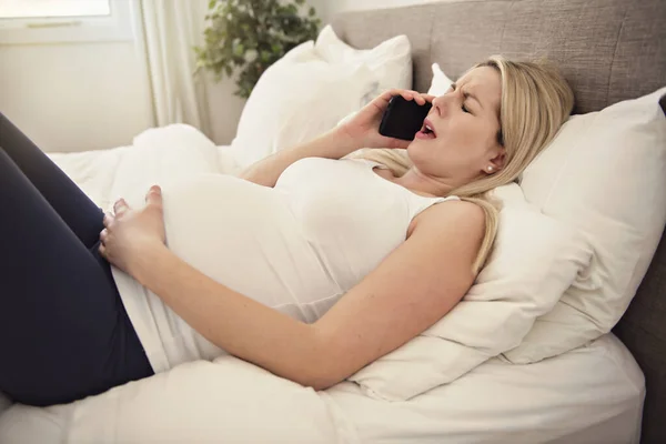 Беременная женщина на кровати дома, у нее схватки с телефоном — стоковое фото