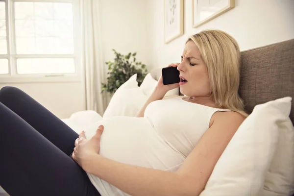 Těhotná žena na posteli doma s kontrakcemi držící mobilní telefon — Stock fotografie