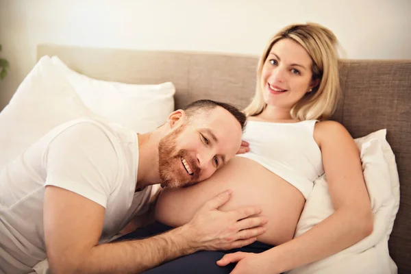 Beautiful pregnant woman and her handsome husband spending time together in bed ear belly — Stock Photo, Image