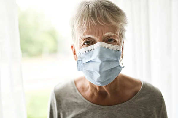 An elderly woman in a protective mask in front of window — Stock Photo, Image