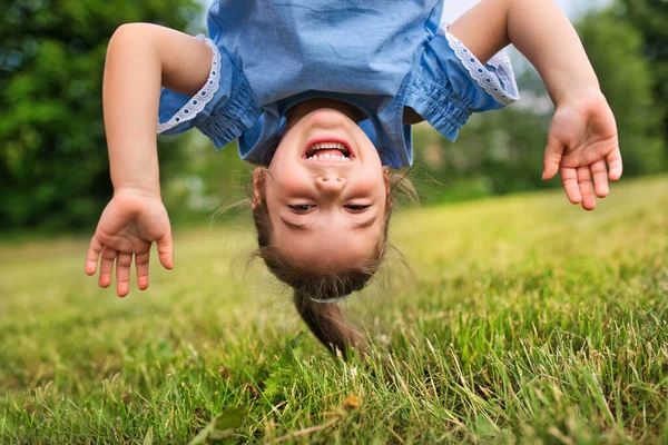 Grappig gelukkig klein meisje dat op haar hoofd staat op het gras in het park. — Stockfoto