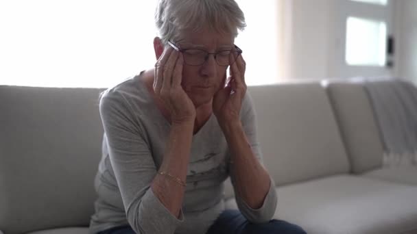 Olycklig huvudvärk Pensionerad äldre kvinna sitter på soffan hemma — Stockvideo