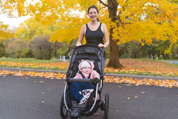 Beautiful young mother with her daughter in jogging stroller outside in autumn nature — Stock Photo, Image