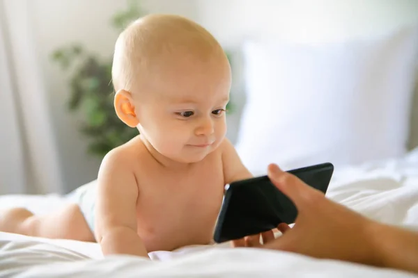 Um retrato de um bebê bonito verificando smartphone deitado em uma cama — Fotografia de Stock