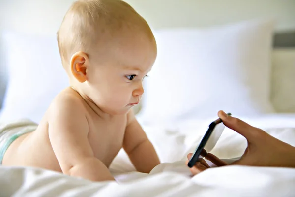 Um retrato de um bebê bonito verificando smartphone deitado em uma cama — Fotografia de Stock