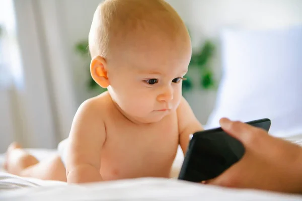 A portrait of a cute baby checking smartphone lying in a bed — Stock Photo, Image