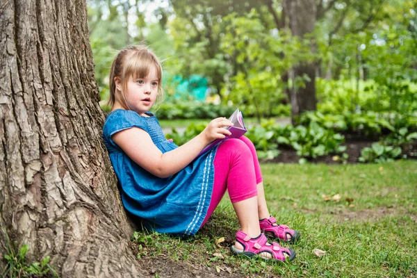 Un portrait de trisomie 21 enfant fille à l'extérieur s'amuser sur un livre de lecture parc — Photo