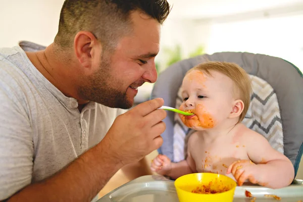 Un pequeño bebé comiendo su cena y haciendo un lío con papá en el lateral — Foto de Stock