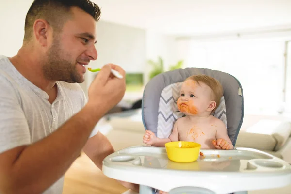 Un pequeño bebé comiendo su cena y haciendo un lío con papá en el lateral — Foto de Stock