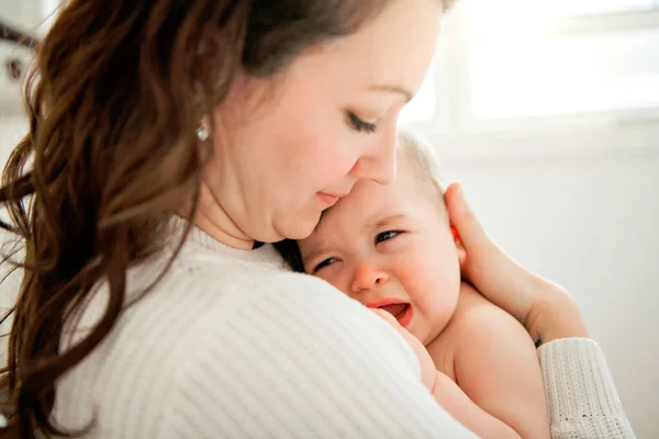 Uma jovem mãe com um bebê em um belo quarto com janela nas costas consolando — Fotografia de Stock