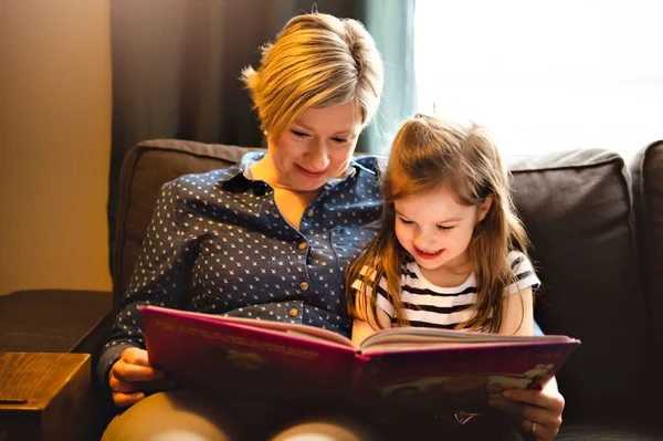 A mother teaching daughter holding book sitting on sofa