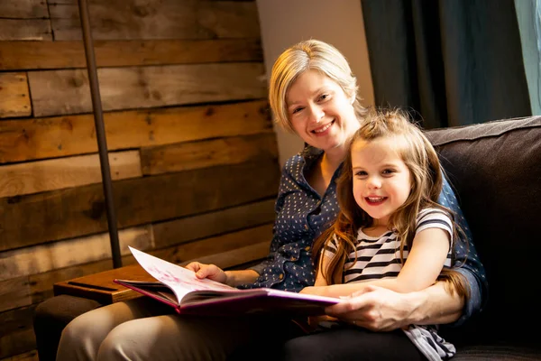 Мать учит дочь держать книгу сидя на диване — стоковое фото