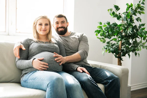 En man omfamnar lycklig gravid kvinna som sitter på soffan — Stockfoto