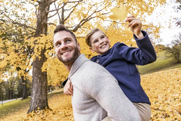 Lycklig familj far och barn pojke i höst löv falla i parken håller sitt barn på ryggen — Stockfoto