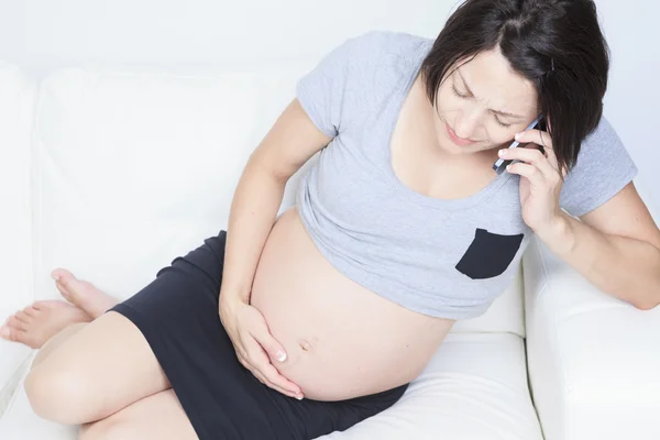 Mulher grávida sentada no sofá com um telefone — Fotografia de Stock