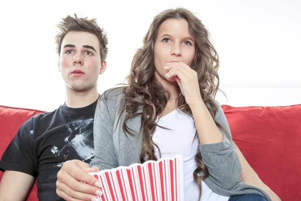 Primer plano retrato de pareja joven sentados juntos en un sofá en casa viendo la televisión, sonriendo alegremente comiendo maíz pop disfrutando de una noche juntos. Hogar estilo de vida y tecnología de entretenimiento . — Foto de Stock