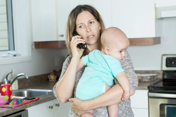 Женщина разговаривает по телефону, держа ребенка на руках на кухне — стоковое фото