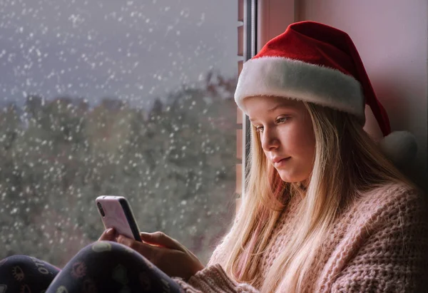Мила дівчина з Санта-Капелюха під час різдвяних свят користується мобільним телефоном і падає сніг за вікном. — стокове фото