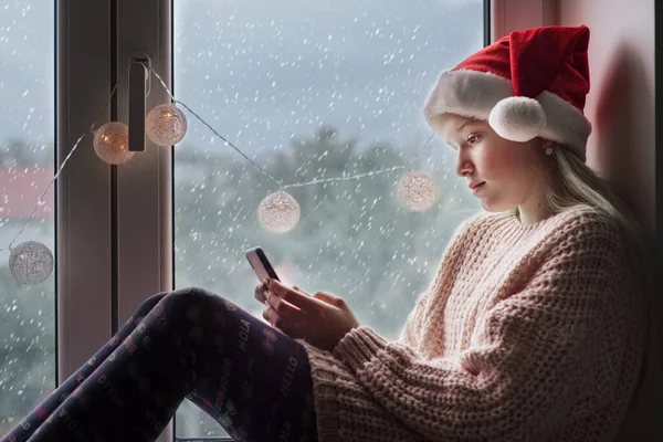 Мила дівчина з Санта-Капелюха під час різдвяних свят користується мобільним телефоном і падає сніг за вікном. — стокове фото