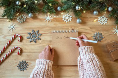 Noel Baba 'ya elinde kalemle gelen mektup ve arka planda Noel ışıkları ve süslemeleri.