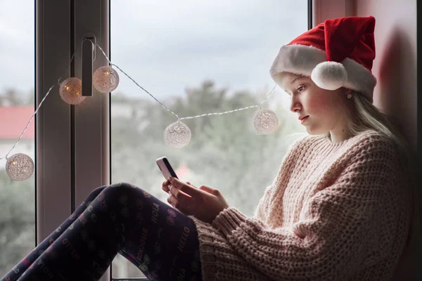 Мила дівчина з Санта - Капелюха під час різдвяних свят користується мобільним телефоном і сидить на підвіконні. — стокове фото