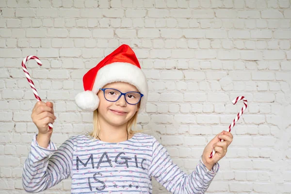 Щаслива маленька дівчинка в капелюсі Санта і окулярах тримає цукерки тростини цукерки на білому тлі стіни — стокове фото