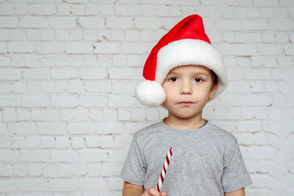 Bonito menino em chapéu de Santa comer doces de Natal cana doces em tijolos brancos parede fundo — Fotografia de Stock