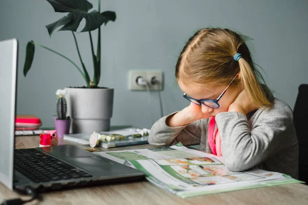 Klein meisje in bril kijkend naar haar klasboek tijdens het hebben van online les thuis — Stockfoto