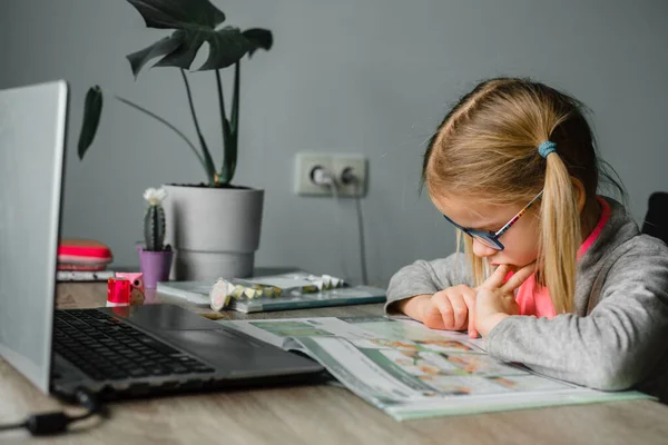 Klein meisje in bril kijkend naar haar klasboek tijdens het hebben van online les thuis — Stockfoto