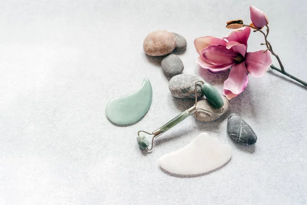 Yüz masajı aletleri: çakıl taşlı yeşim guasha masaj silindiri ve çakıl taşlı kazıyıcı ve açık gri arka planda mor manolya çiçeği, SPA rahatlama, yüz ve cilt bakımı