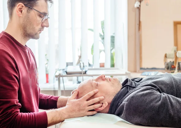 CST terapisti, erkek bir hastanın kafatası, osteopati ve manuel terapi eklemlerini elle muayene etmek için nazik dokunuşlar kullanır.