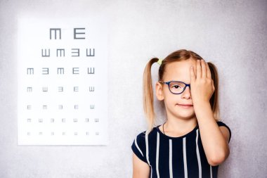 Little girl wearing eyeglasses covering her eye with one hand taking eyesight test before school with blurry eye chart at the background clipart
