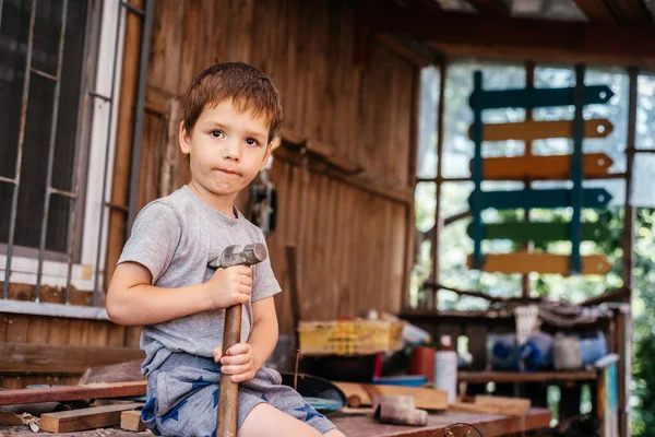 Pequeño niño martillos clavos con un martillo en una tabla de madera en el taller de carpinteros — Foto de Stock