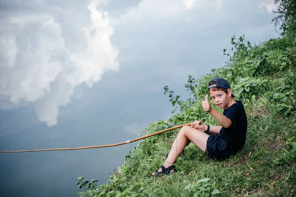 Un niño sosteniendo una caña de pescar con el pulgar hacia arriba, pesca en el lago, actividades de verano y pasatiempos para los niños — Foto de Stock