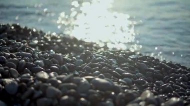 Işıl ışıl güneşin ön planında çakıl ve kayalar sakin deniz yüzeyinde batıyor.