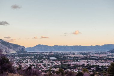 Sicilya, Palermo 'nun yükseltilmiş panoramik manzarası, gün batımında yumuşak bir gökyüzü, arka planda dağlar ve aşağıda yoğun şehir binaları. Seyahat, kentsel ve peyzaj temaları için ideal