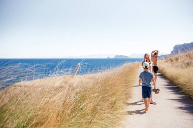 Üç çocuk Sicilya kıyı şeridi boyunca, uzun otlar ve deniz manzaralarıyla çevrili bir yolda yalınayak yürüyor. Aile, seyahat, yaz ve çocukluk özgürlüğü temaları için ideal