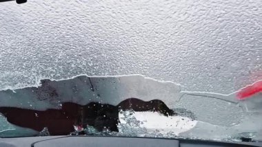 Kışlık ceket giyen bir adam, aracın içinden görünen bir buz kazıyıcı kullanarak arabanın ön camından kalın buzlar kazıyor. Sahne, dondurucu yağmur veya soğuk hava koşulları sonrasında ortak bir kış rutinini yakalıyor.