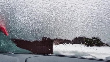 Kışlık ceket giyen bir adam, aracın içinden görünen bir buz kazıyıcı kullanarak arabanın ön camından kalın buzlar kazıyor. Sahne, dondurucu yağmur veya soğuk hava koşulları sonrasında ortak bir kış rutinini yakalıyor.