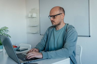Ciddi orta yaşlı, gözlüklü bir adam ev ofisteki bir masada dizüstü bilgisayarda yazı yazıyor. Konsantrasyonunu, kararlılığını ve uzak çalışma ve profesyonel sorumlulukla ilgili hafif stresi ifade ediyor.