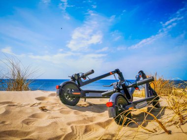 Denizin kenarındaki kum tepelerinde duran iki elektrikli scooter.
