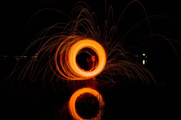 Yanan Çelik Yün kıvrımları, yanan kıvılcımların yörüngeleri bahar gecesi manzarasının arka planında su gölü yüzeyinde.